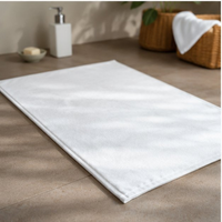 White bath mat on a tiled bathroom floor with a basket and soap dispenser in the background.